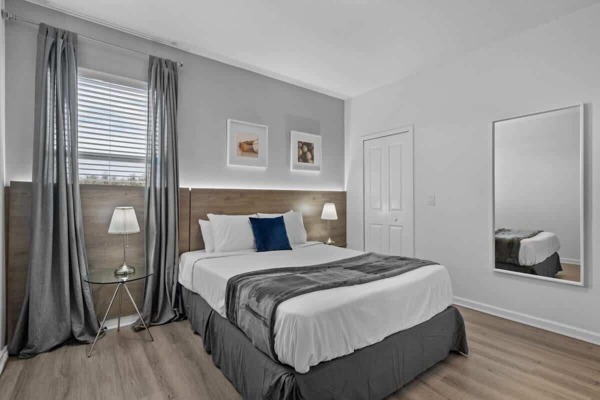 Una habitación de hotel con una cama grande, una mesita de noche con lámpara, una ventana con cortinas y un espejo adosado a la pared.