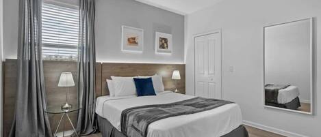 Una habitación de hotel con una cama grande, una mesita de noche con lámpara, una ventana con cortinas y un espejo adosado a la pared.