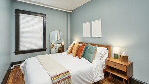 A bedroom with a bed, bedside table, lamp, and a window with blinds. - The Inn Too @ The Highland Hill (Akron)