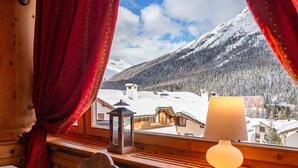 Una habitación con detalles de madera, una ventana con cortinas rojas, una lámpara de mesa y vistas a casas y montañas nevadas.