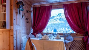 Un comedor con una mesa dispuesta para dos, sillas de madera, un ventanal con cortinas rojas y vistas a un paisaje nevado.