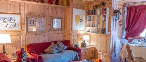 A cozy wooden cabin interior with a red sofa, a blue armchair, and a dining area with a table and chairs.