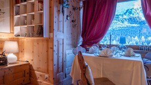 Un comedor acogedor con una mesa de madera dispuesta para dos personas, una estantería y una ventana que ofrece vistas a un paisaje nevado.
