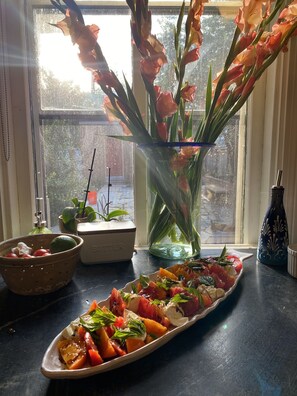 Una mesa con una ensalada de frutas, un florero con flores y una ventana con vista a árboles.