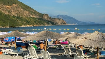 Une plage avec des chaises longues blanches et des parasols en chaume, une colline rocheuse en arrière-plan et une mer calme.