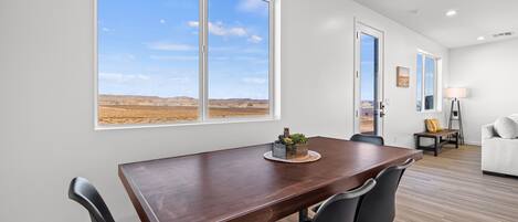 A dining area with a wooden table and black chairs, a large window with a desert view, and a white sofa in the background.