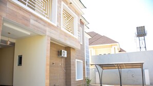 Une maison moderne à deux étages, dotée d’un espace extérieur aménagé avec un coin salon couvert, d’un petit jardin et d’un tuyau d’arrosage.