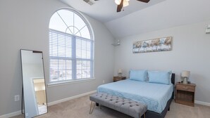Ein Schlafzimmer mit großem Fenster, Deckenventilator, Bett mit blauer Bettwäsche, einer Bank und einem Nachttisch mit Lampe.