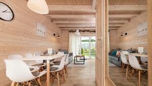 Un interior moderno con paredes de madera, un área de comedor con sillas blancas, un sofá y vistas a un jardín a través de un ventanal.