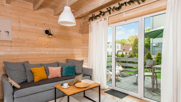 Una habitación acogedora con un sofá gris, una mesa de centro de madera y una computadora portátil. Hay un ventanal con vistas a un jardín y edificios.