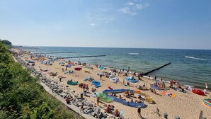 Una playa con un muelle largo que se adentra en el mar, zonas arenosas con sombrillas y gente disfrutando, y un cielo despejado.