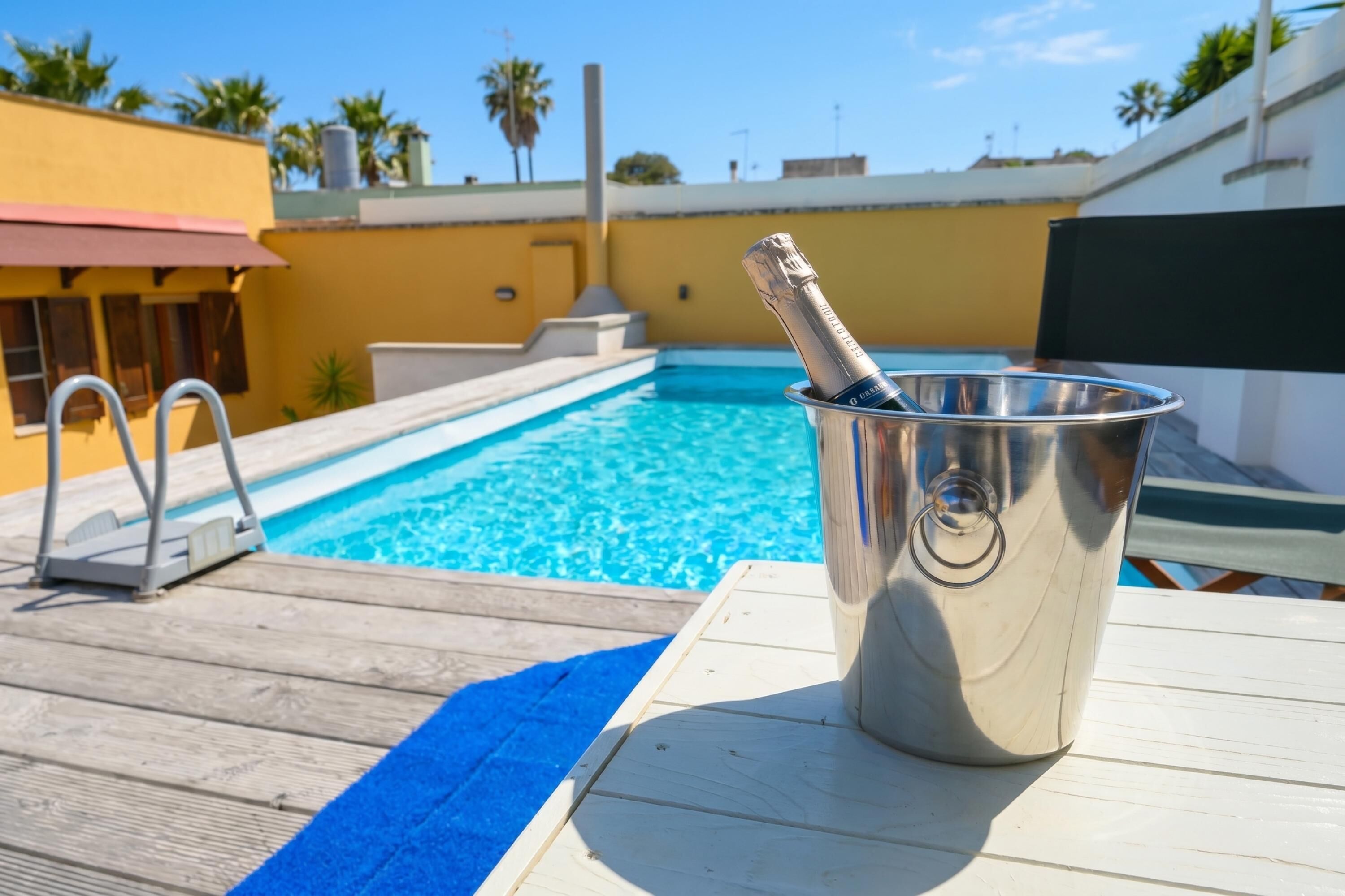 A poolside area with a metal bucket containing a bottle, a wooden deck, and a clear blue pool.