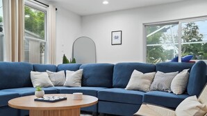 A modern living room with a blue sectional sofa, a wooden coffee table, and a view of greenery outside. - 4BR Heated Pool|12min to Beach|Gameroom|King beds (Clearwater)