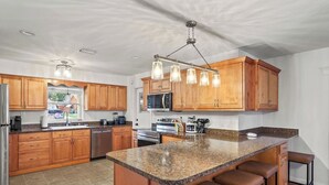 A modern kitchen with wooden cabinets, granite countertops, a central island with bar stools, and a hanging light fixture. - 4BR Heated Pool|12min to Beach|Gameroom|King beds (Clearwater)
