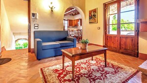 A living room with a blue sofa, a wooden coffee table, a red and white patterned rug, a wooden door, and a view of a kitchen through an archway. - Fratta Orchidee, Cortona, Italy (Cortona)