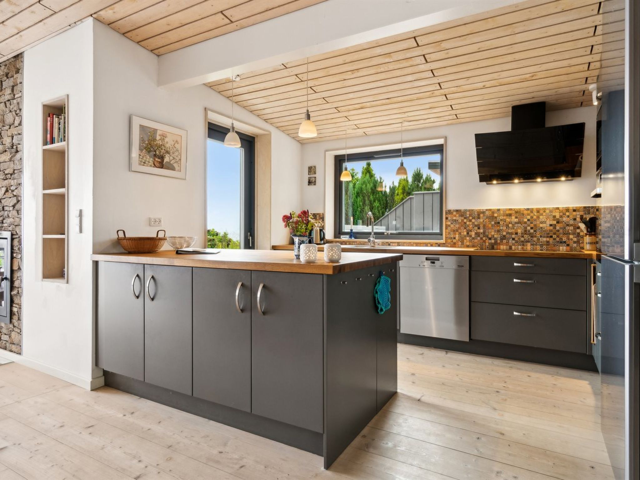 A modern kitchen with a central island, grey cabinetry, a stone accent wall, and a wooden ceiling.