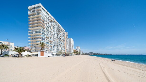 Un hotel frente a la playa con varios pisos, una playa de arena y un cielo azul despejado.