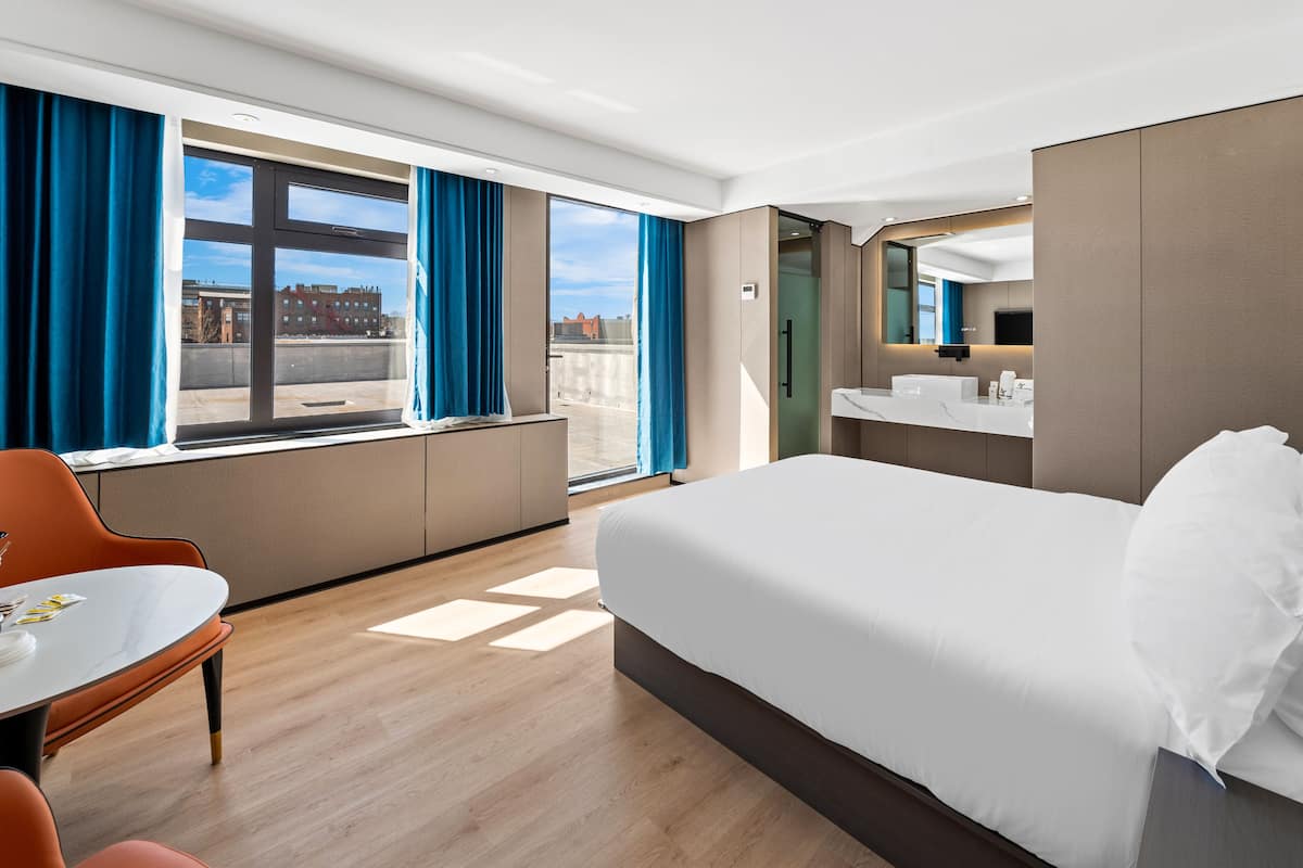Une chambre d’hôtel avec un grand lit, un bureau, une chaise, une salle de bain avec un miroir et une vue sur les bâtiments par la fenêtre.