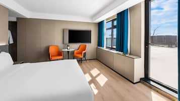 A modern hotel room with a large bed, two orange chairs, a small table, a TV, and a large window with curtains.