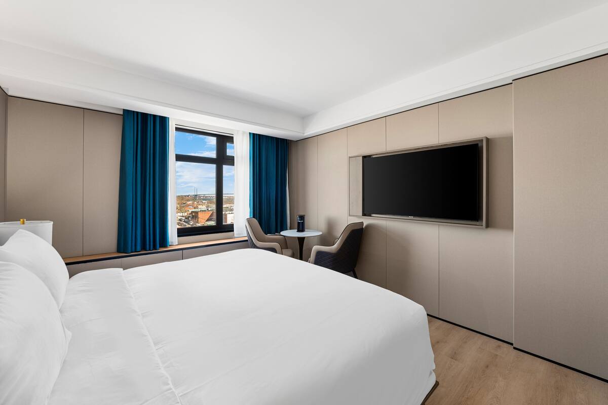 Une chambre d’hôtel avec un grand lit, un téléviseur fixé au mur, une petite table avec des chaises et une fenêtre avec des rideaux.