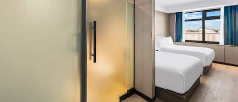 A hotel room with a sliding door, two beds, a large window with curtains, and a view of the outside.