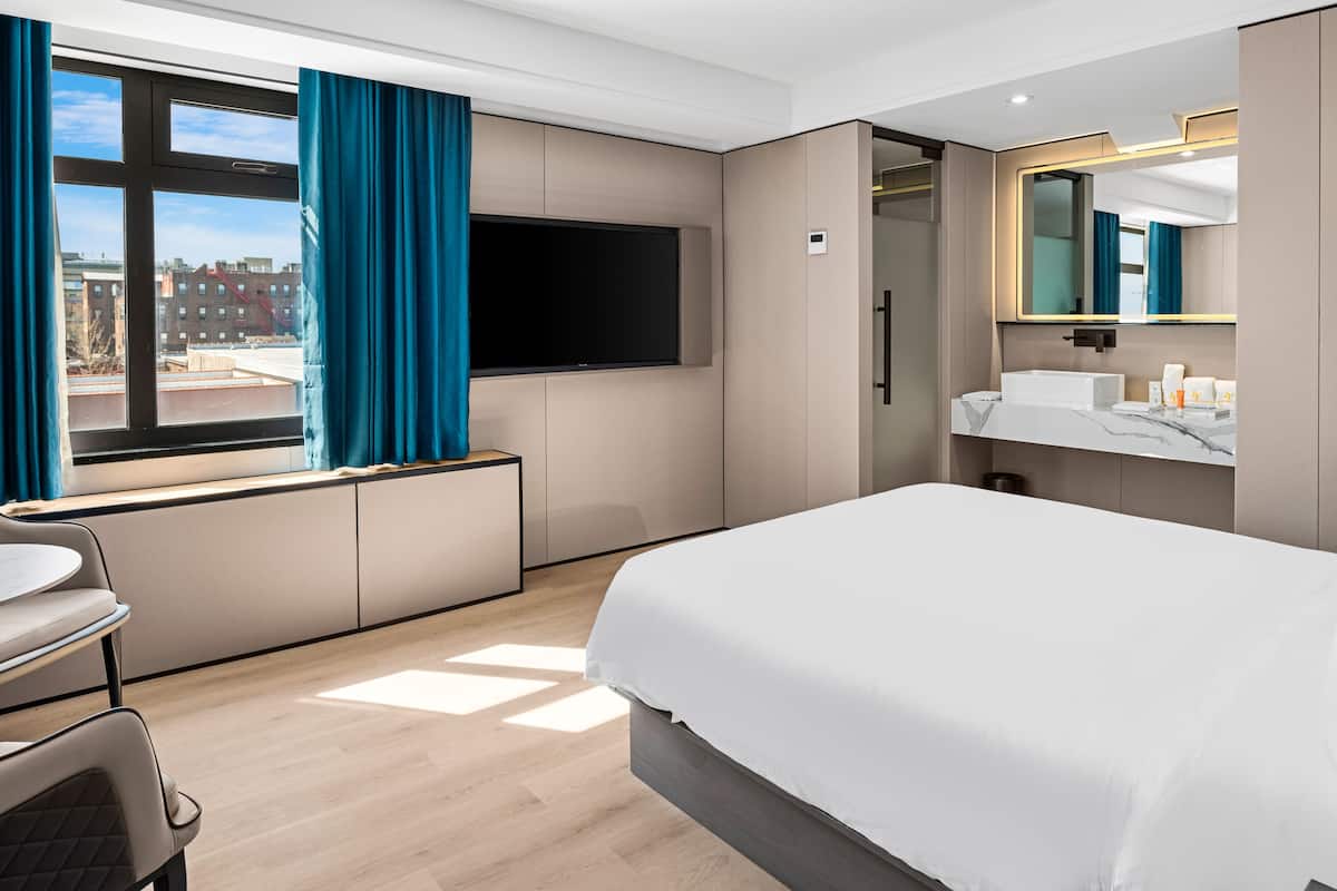 Une chambre d’hôtel moderne dotée d’un grand lit, d’un téléviseur à écran plat fixé au mur, d’une salle de bain avec un lavabo et un miroir, et d’une fenêtre avec des rideaux.