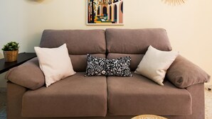 A brown sofa with three cushions, a framed cityscape poster, a woven sun decoration, and a small potted plant.