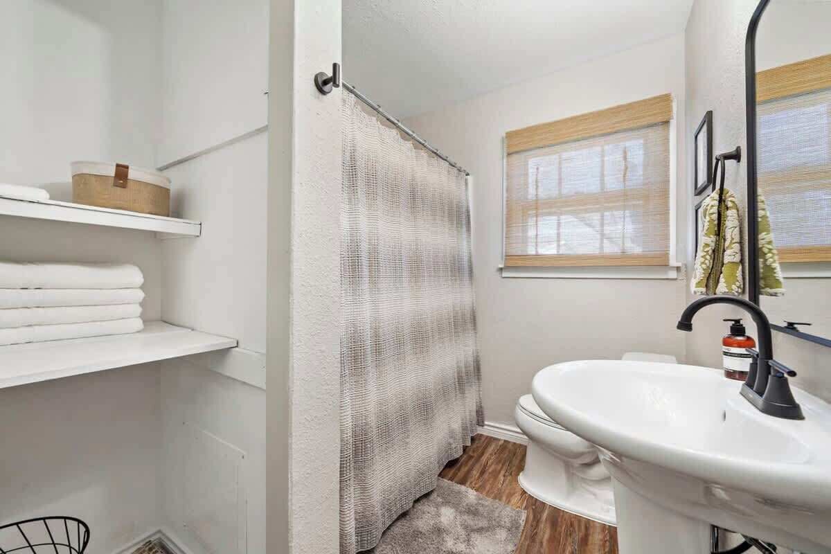 Une salle de bain avec un lavabo blanc, des toilettes, un rideau de douche et une fenêtre avec des stores.