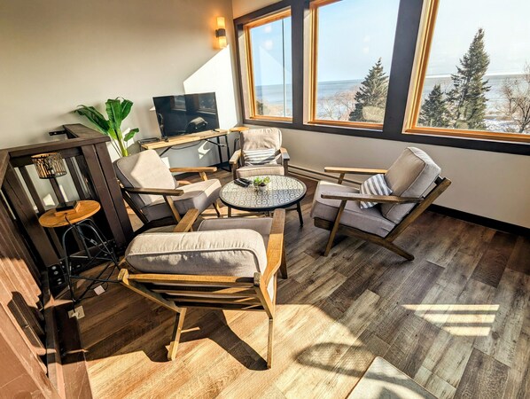 A cozy room with a wooden floor, a round table with chairs, a TV, and a large window with a view of trees and the sea.