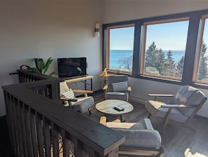 Een moderne woonkamer met een televisie, zitplaatsen en een groot raam met uitzicht op zee.