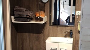 Une petite salle de bain avec des toilettes blanches, un lavabo avec un miroir et une étagère où sont pliées des serviettes.