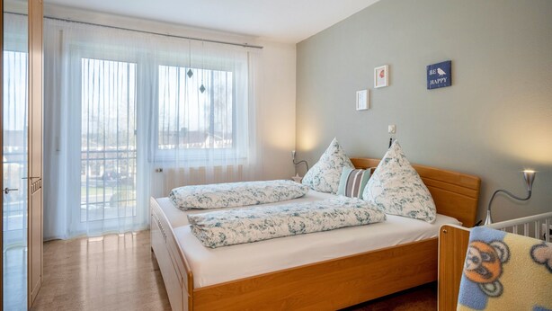 Ein Schlafzimmer mit einem hölzernen Bett, einem Fenster mit transparenten Vorhängen und einer Wand mit der Aufschrift 'BE HAPPY'.