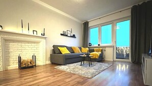 A living room with a fireplace, a sofa with yellow cushions, a coffee table, and a large window with curtains.