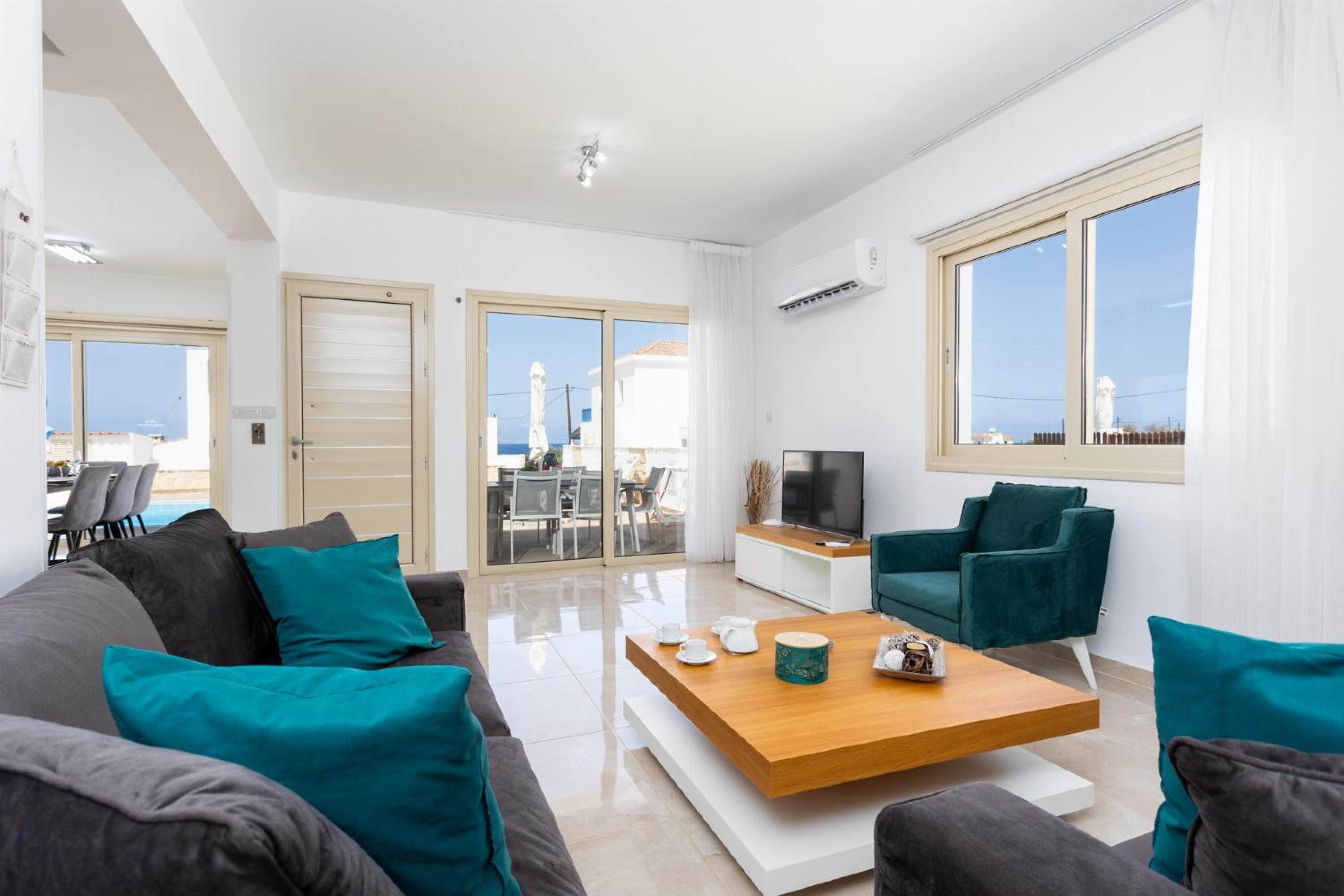 Un salon moderne avec une table basse en bois, des canapés turquoise et gris, un téléviseur et une grande fenêtre donnant sur la mer.