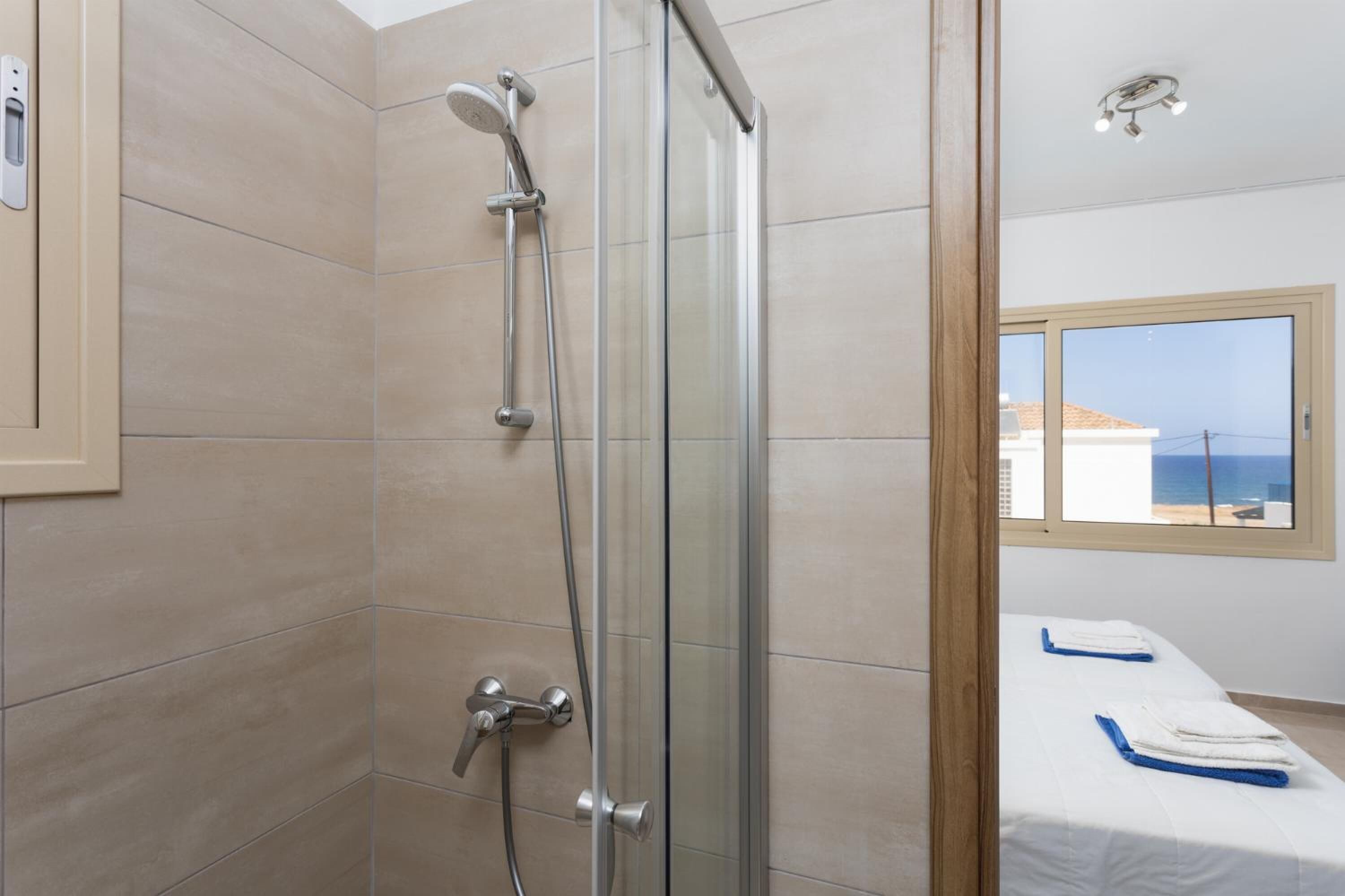 A modern bathroom with a glass-enclosed shower, a small bed with a view of the sea, and a window with a view of the beach.