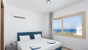 Un dormitorio con dos camas, un equipo de aire acondicionado, una mesita de noche con lámpara y una ventana con vista al mar.