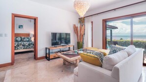 Ein modernes Wohnzimmer mit Fernseher, Sofa, Couchtisch und großen Fenstern mit Blick auf das Meer.