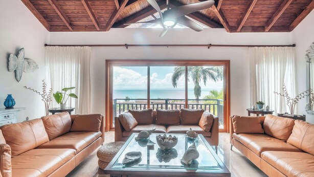 Ein Wohnzimmer mit einem Glastisch, zwei Ledersofas, einem Deckenventilator und Blick auf den Ozean.