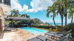 Ein Swimmingpool mit Liegestühlen, einer überdachten Terrasse und Blick auf den Ozean.