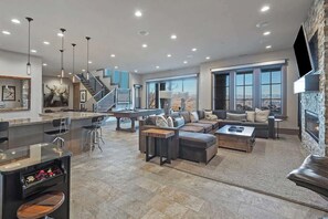 A spacious living area with a large sofa, a coffee table, and a fireplace. There is a kitchen area with a bar and a pool table.