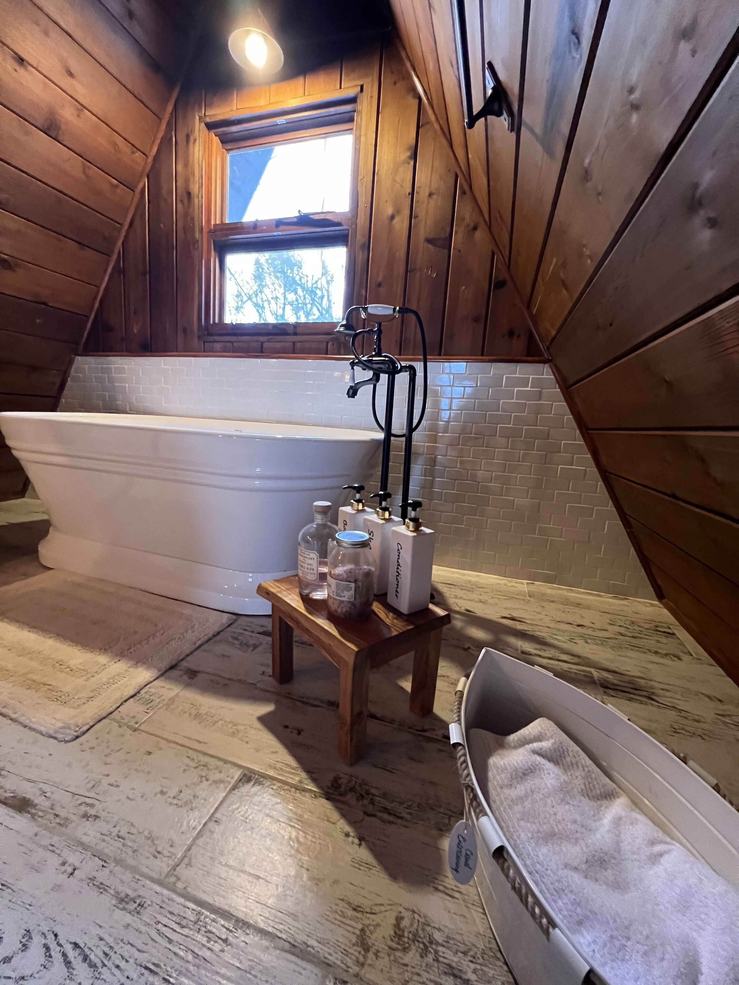 Un baño de madera con una bañera independiente, un taburete de madera pequeño con artículos de aseo, y un ventanal con vistas a árboles.