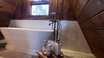 Un baño de madera con una bañera independiente, un taburete de madera pequeño con artículos de aseo, y un ventanal con vistas a árboles.