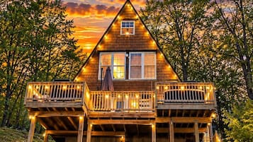 A wooden cabin with a balcony, surrounded by trees, and a sunset in the background.