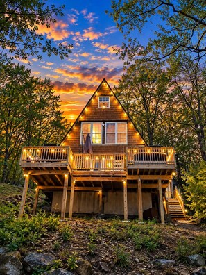 Une cabane en bois dotée d’un balcon, entourée d’arbres, avec un coucher de soleil en arrière-plan.