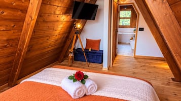 A cozy attic bedroom with a wooden sloped ceiling, a flat-screen TV mounted on the wall, a bed with an orange quilt, and a small window.