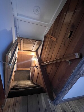 Una escalera de madera con barandilla, una lámpara en el techo y una puerta con un pequeño ventanal.