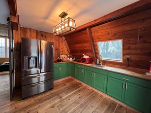 Una cocina con paredes de madera, gabinetes verdes, un refrigerador de acero inoxidable y una ventana con vista a árboles.