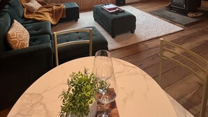 Ein gemütliches Dachzimmer mit Holzbalkendecke, Kamin, einer Couch und einem Esstisch mit Pflanze und Weinglas.