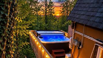 Un bain à remous sur une terrasse en bois, illuminé de guirlandes lumineuses, entouré d’arbres et avec un coucher de soleil en arrière-plan.