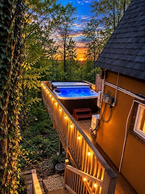 A hot tub on a wooden deck with string lights, surrounded by trees and a sunset in the background.